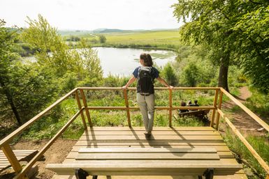 La randonneuse se tient sur une plateforme en bois avec un banc et regarde par-dessus la balustrade le Eichholzmaar, qui se trouve au milieu d'un paysage verdoyant.
