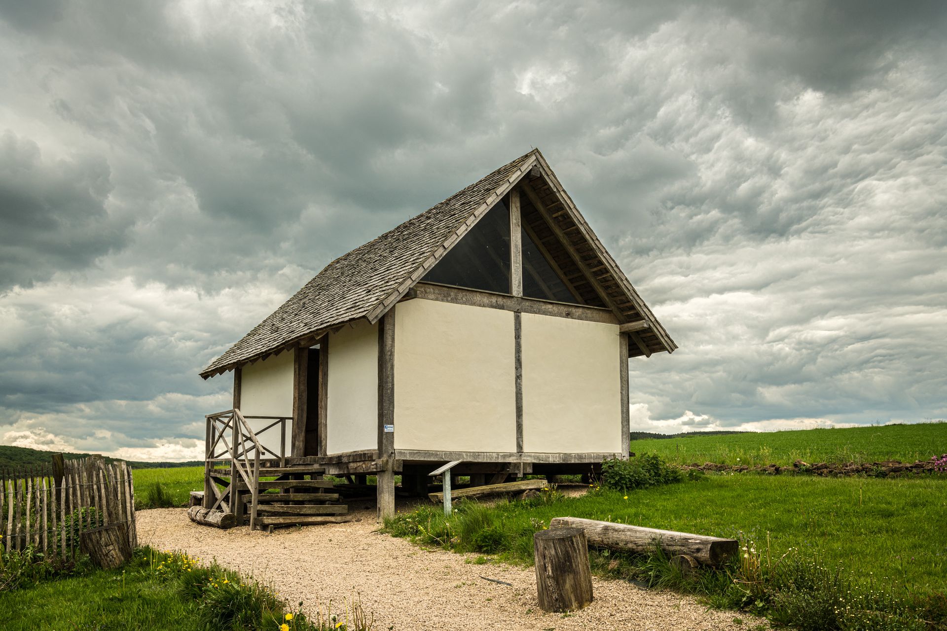 Weg zu einem Holzhäuschen auf einer grünen Wiese, bewölkter Himmel im Hintergrund.