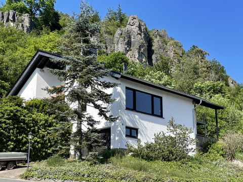 Une maison moderne entourée d'une verdure luxuriante et des rochers en arrière-plan. Le ciel est bleu et clair.