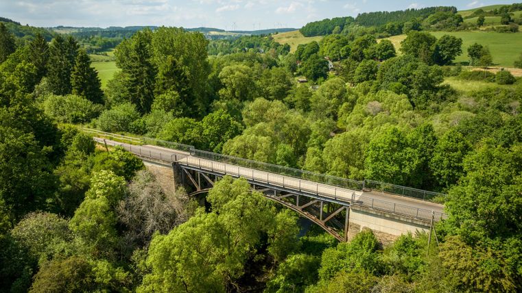 Luchtaufnahme van de Kyll-fietspadbrug in Stadtkyll, omringd door weelderige groene vegetatie en zachte heuvels onder een blauwe lucht.
