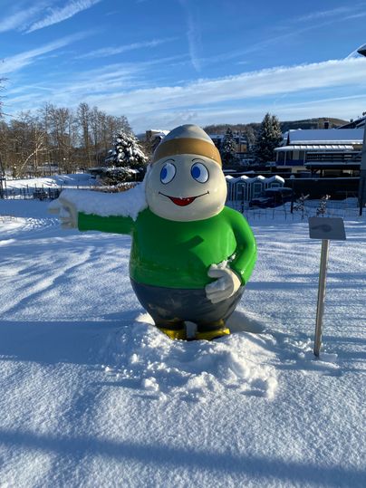 Statue von Maskottchen Willi Basalt steht in einem mit viel Schnee bedecktem Park und zeigt nach links. Hinter ihm der Park und strahlend blauer Himmel.