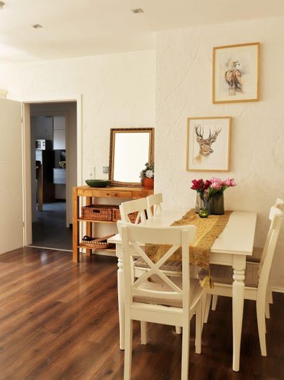 Een lichte eetkamer met een witte tafel en stoelen. Aan de muur hangen schilderijen, en op de tafel staan bloemen.