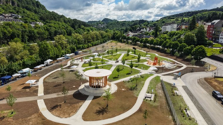 Luchtfoto van een park met paden, bomen en een paviljoen, omringd door heuvels en huizen.