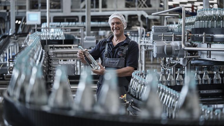 Ein Arbeiter in einer Abfüllanlage überprüft eine Wasserflasche. Er trägt Arbeitskleidung und eine Haube. Im Hintergrund sind viele Flaschen auf einem Förderband.