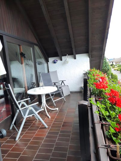 Ein Balkon mit zwei Liegestühlen und einem kleinen Tisch. Bunte Geranien blühen in Kästen entlang der Geländer.