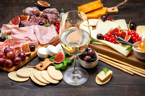 Auf einem dunklen Holzbrett liegen diverse Antipasti (Schinken, Salami, Käse, aufgeschnittene Feigen, Trauben, Cracker) und ein Glas Wein