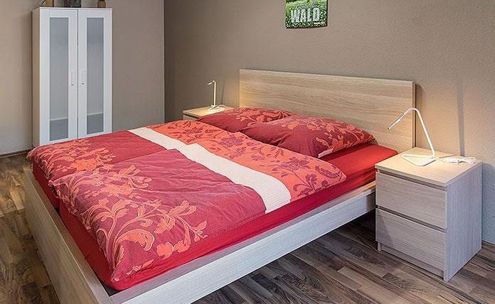 Een gezellige slaapkamer met een houten bed en rode beddengoed. Op de achtergrond staan een kast en een nachttafeltje met een lamp.