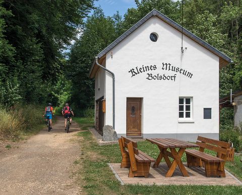 Weißes Gebäude mit der Aufschrift 'Kleines Museum Bolsdorf'. Zwei Radfahrer auf einem Weg daneben. Holztisch und Bänke im Vordergrund.