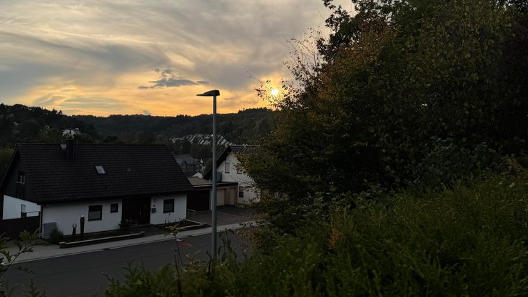 Une vue tranquille du soir avec des maisons et des arbres au premier plan. Le soleil se couche derrière les collines et colore doucement le ciel.