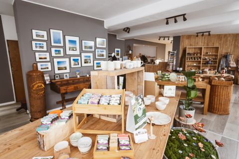 L'image montre un magasin avec des savons faits à la main, des articles en céramique et des décorations, exposés sur des tables en bois et des étagères. Des photographies encadrées de paysages sont accrochées au mur.