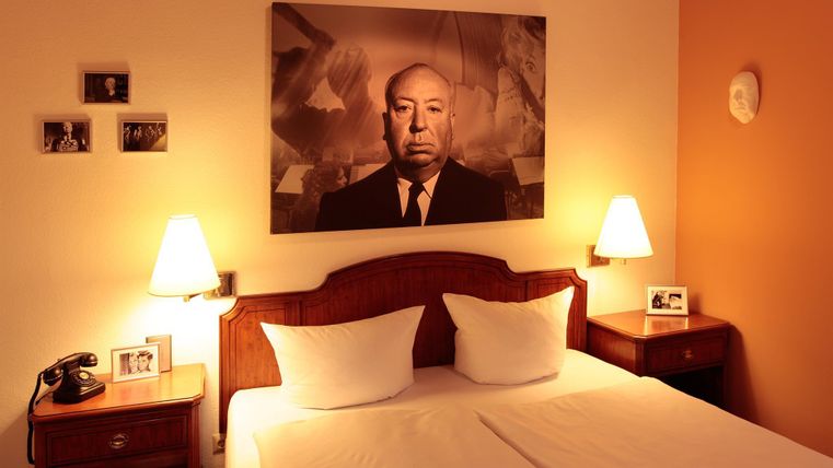 Une chambre confortable avec un grand lit et deux coussins.
Sur le mur, un portrait d'un homme est accroché, et sur la table de nuit se trouvent des photos et un téléphone.
