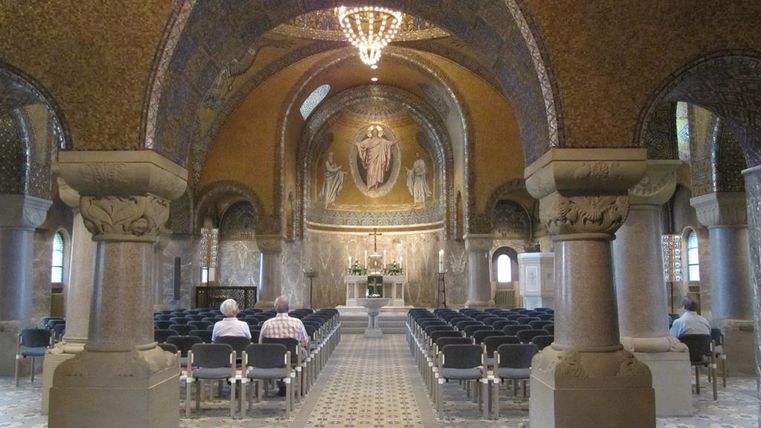 Ein schöner, ruhiger Innenraum einer Kirche mit goldenen Mosaiken. Die Sitzplätze sind ordentlich angeordnet und eine große Deckenleuchte bringt Licht in den Raum.