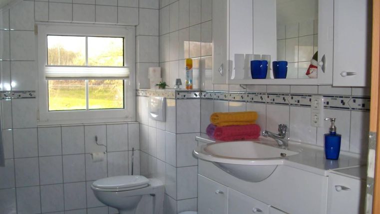 Ein modernes Badezimmer mit weißen Fliesen und einem großen Fenster. Auf dem Waschbecken liegen bunte Handtücher und es gibt verschiedene Pflegeprodukte.