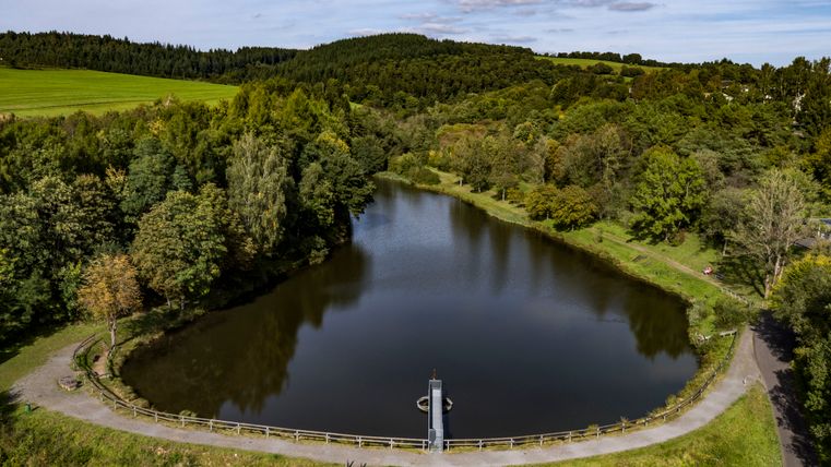 View of the Gerolstein reservoir from above, surrounded by forests and meadows. The dark blue lake stands out against the green landscape.