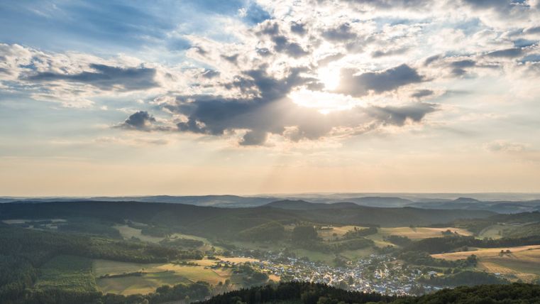 Vue de paysage de Neroth avec des collines boisées et un village dans la vallée, sous un ciel de nuages et de rayons de soleil.