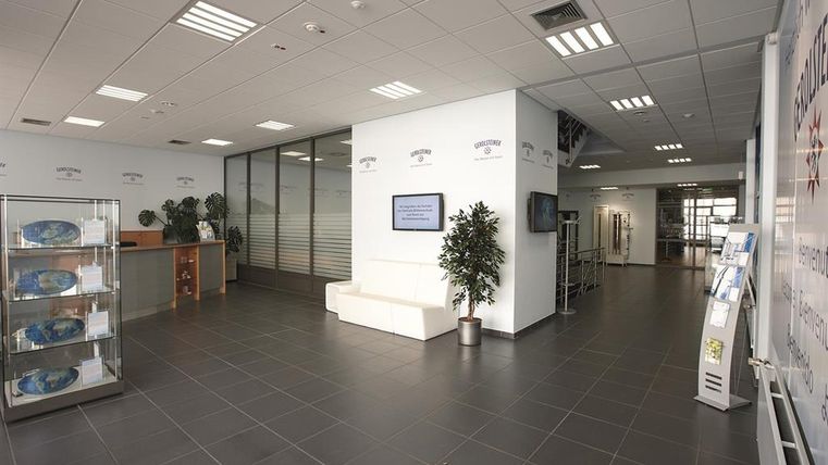 Empfangsbereich im Gerolsteiner Besucherzentrum mit Tresen, Sitzgelegenheiten und Pflanzen. An der Wand ein Bildschirm mit Begrüßungstext.