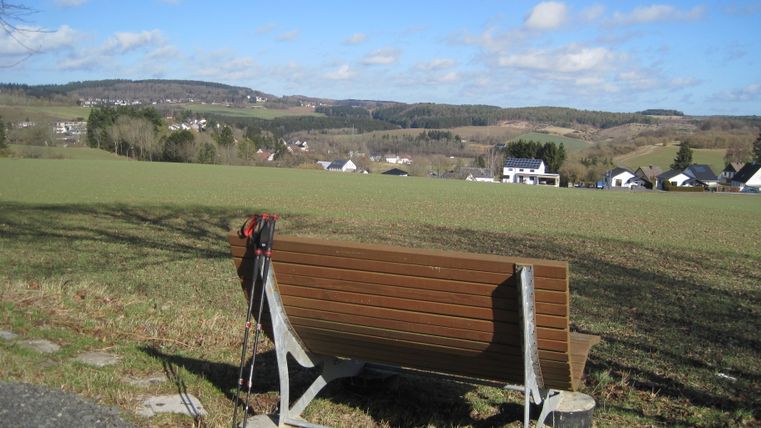 Bank mit Wanderstöcken vor einer ländlichen Landschaft mit Hügeln und Häusern.