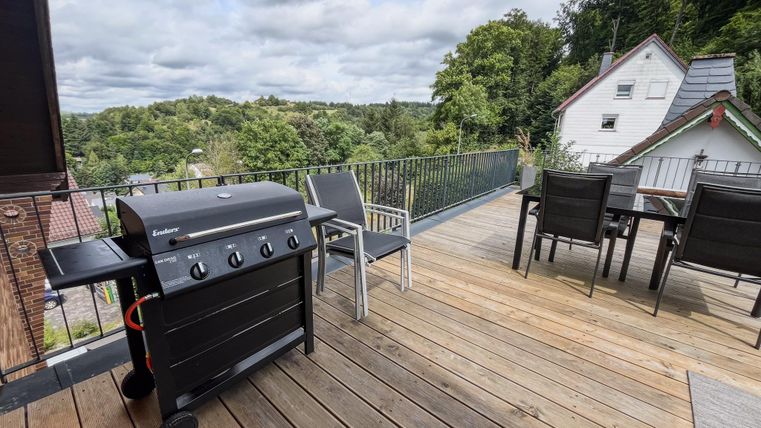 Eine große Terrasse mit einem Grill und mehreren Sitzgelegenheiten. Im Hintergrund sind Wald und moderne Häuser zu sehen.