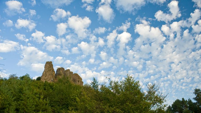 A rocky elevation surrounded by trees and a clear blue sky. White, fluffy clouds drift across the sky.