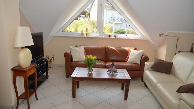 Een gezellige woonkamer met een bruin leren bank en een witte bank. Een houten tafel en een tv-console completeren de inrichting, terwijl grote ramen veel licht binnenlaten.