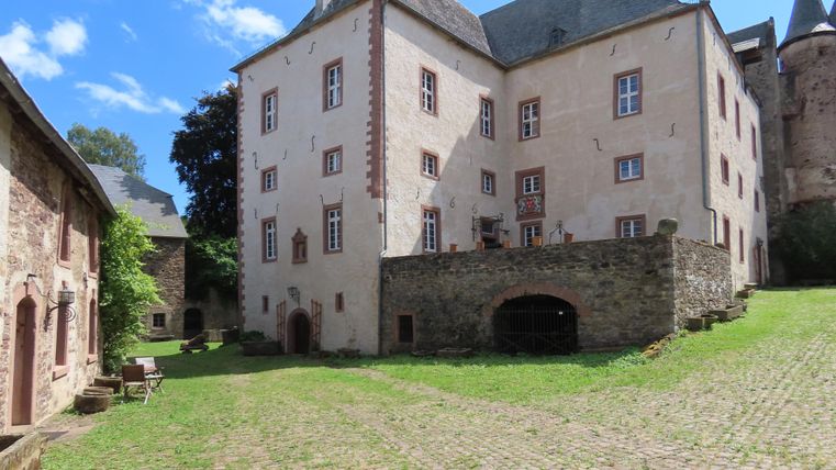 Die Aufnahme zeigt eine alte Burg oder ein Herrenhaus mit einem Steingebäude im Vordergrund und einem weiteren Gebäude aus Stein und Holz im Hintergrund. Die Gebäude sind von Grünflächen umgeben.