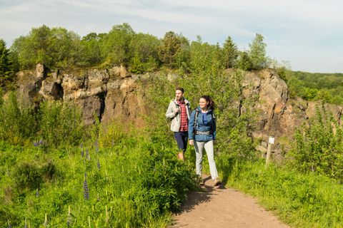 Zwei Wanderer im Vulkangarten Steffeln vor der großen Felswand des Vulkans Steffelberg umgeben von grünen Sträuchern und Bäumen.