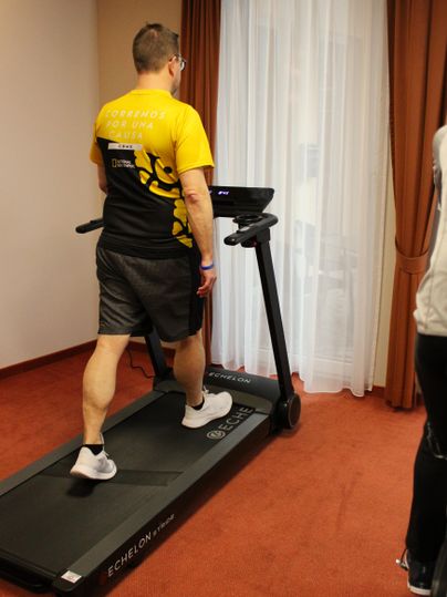 Un homme court sur un tapis roulant dans une pièce lumineuse. Il porte un t-shirt jaune et un short de sport.