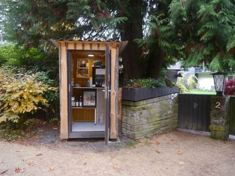 Kleiner Holzschuppen mit Produkten im Inneren, umgeben von Bäumen und einem Gartenzaun mit der Nummer 2.