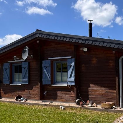 Ein gemütliches Holzhaus mit blauen Fensterläden. Der Garten ist grün und ein sonniger Himmel ist im Hintergrund zu sehen.