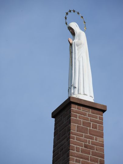 Eine Statue der Jungfrau Maria steht auf einem Ziegelturm. Der Himmel ist klar und blau.