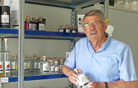 Ein älterer Mann steht vor einem Regal mit verschiedenen Flaschen. Er hält eine Flasche in der Hand und schaut freundlich in die Kamera.
