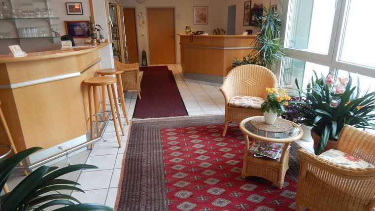 Ein einladender Empfangsbereich mit einer Holztheke und gemütlichen Rattanmöbeln. Grüner Pflanzenschmuck und ein roter Teppich verleihen dem Raum eine warme Atmosphäre.