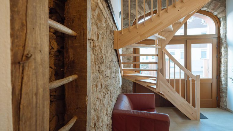 Ein heller Flur mit einer Holztreppe und einem roten Sofa. Die Wände bestehen aus Stein, was dem Raum einen rustikalen Charme verleiht.