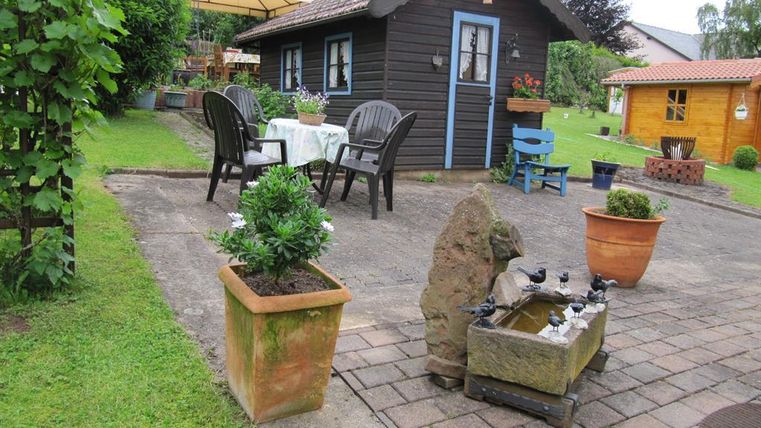 Ein gemütlicher Garten mit einem Holzhaus und einem kleinen Tisch. Töpfe mit Pflanzen und eine gemütliche Sitzgruppe laden zum Entspannen ein.