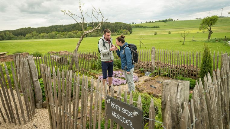 Zwei Personen stehen in einem umzäunten Kräutergarten auf dem Land. Ein Schild weist auf "Römische Kräuter" hin. Im Hintergrund sind Felder und Bäume zu sehen.