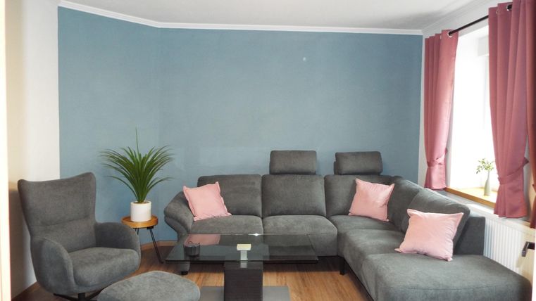 Ein modernes Wohnzimmer mit einem grauen Sofa und einem gemütlichen Sessel. An den Wänden sind hellblaue und rosa Akzente, sowie eine Pflanze auf dem Tisch.