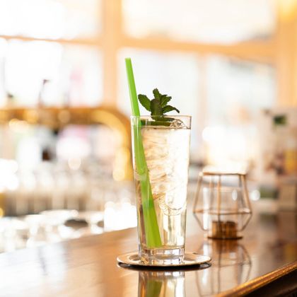 A refreshing drink in a tall glass with ice, a green straw, and a mint leaf. The background features a stylish bar atmosphere.