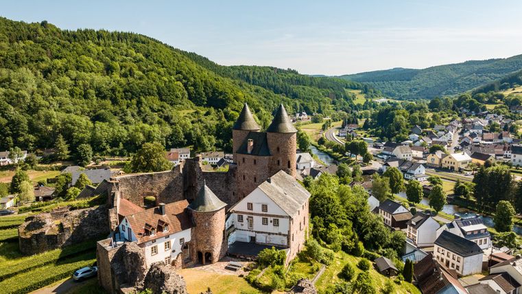 Luftaufnahme der Bertradaburg in Mürlenbach, umgeben von grünen Hügeln und dem Dorf. Die Burg hat markante Türme und liegt in einer idyllischen Landschaft.