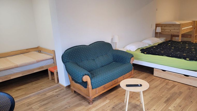 Une chambre simple avec un canapé bleu, une petite table et deux lits. Le sol est en bois et la pièce semble lumineuse et accueillante.