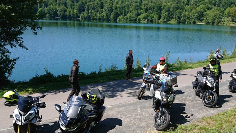 Un groupe de motards se tient au bord d'un lac tranquille. En arrière-plan, on aperçoit de grands arbres verts.
