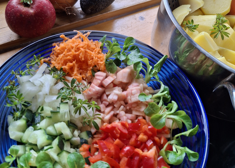 Auf einem Holzbrett steht ein Teller mit einem Salat mit Gurken, Paprika, Zwielbeln, Karotten und Fisch; daneben steht ein Topf mit Kartoffeln