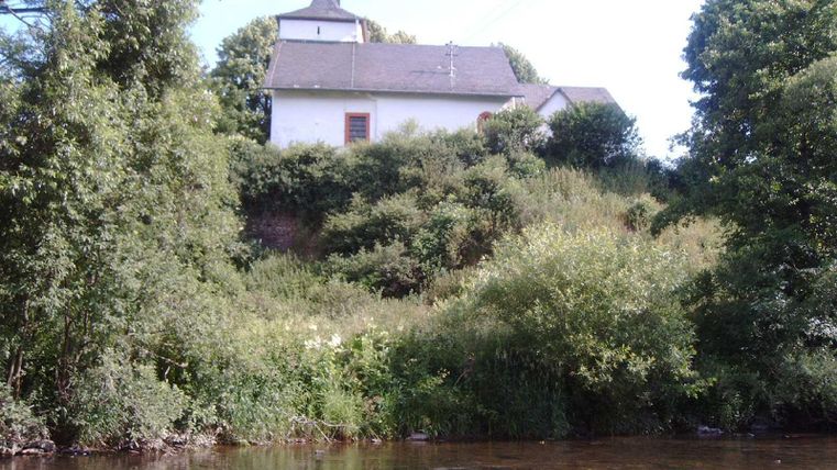 Eine Kirche steht auf einem bewachsenen Hügel, umgeben von Bäumen. Im Vordergrund fließt ein ruhiger Fluss.