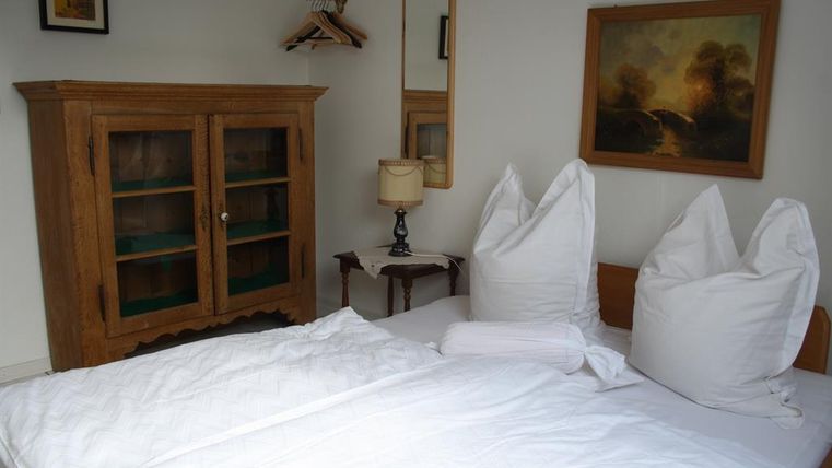 Ein einfaches Schlafzimmer mit einem Bett, zwei großen Kissen und einer weißen Bettdecke. Im Hintergrund stehen ein Holzschrank und ein Tisch mit einer Lampe.