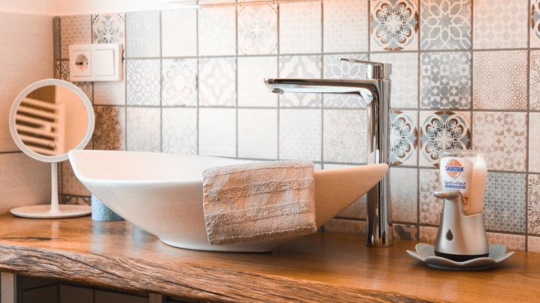 Ein modernes Badezimmer mit einem eleganten Waschbecken und einem Holzregal. Ein Spiegel und Pflegeprodukte vervollständigen die ansprechende Einrichtung.