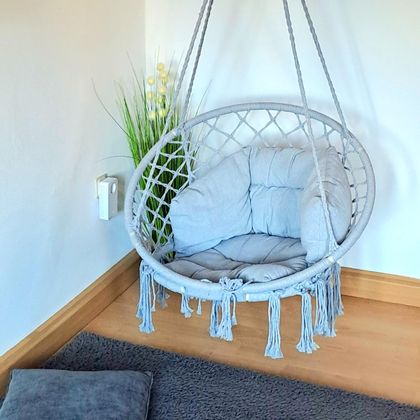 Ein stylischer Hängehocker in Grau mit gemütlichen Kissen. Im Hintergrund sind grüne Pflanzen und ein weicher Teppich zu sehen.