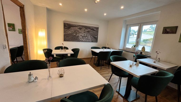 Ein elegantes Café mit hellen Tischen und grünen Stühlen. Der Raum ist freundlich und modern gestaltet, mit großen Fenstern und sanfter Beleuchtung.