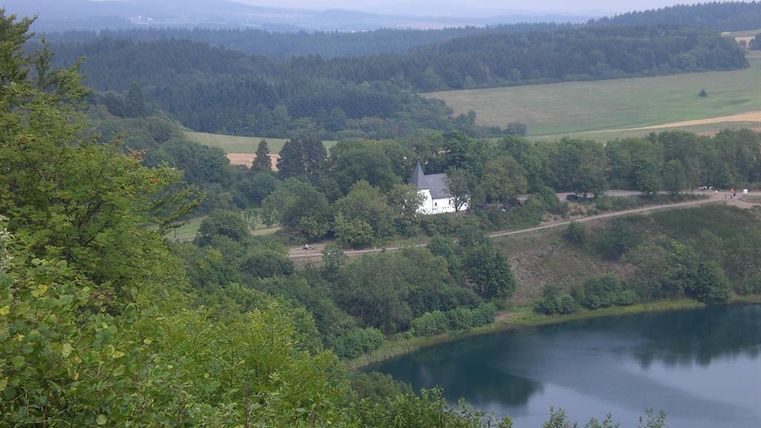 Eine ruhige Landschaft mit einem kleinen Gebäude, umgeben von Bäumen. Im Vordergrund befindet sich ein klarer See, der die grüne Umgebung reflektiert.