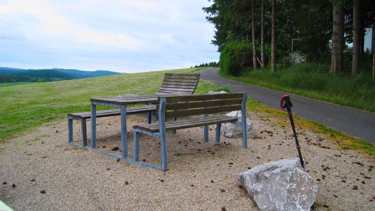 Sitzbank und Tisch auf Kiesfläche mit Wanderstock, umgeben von Wiesen und Bäumen. Ein Weg führt durch die Landschaft.