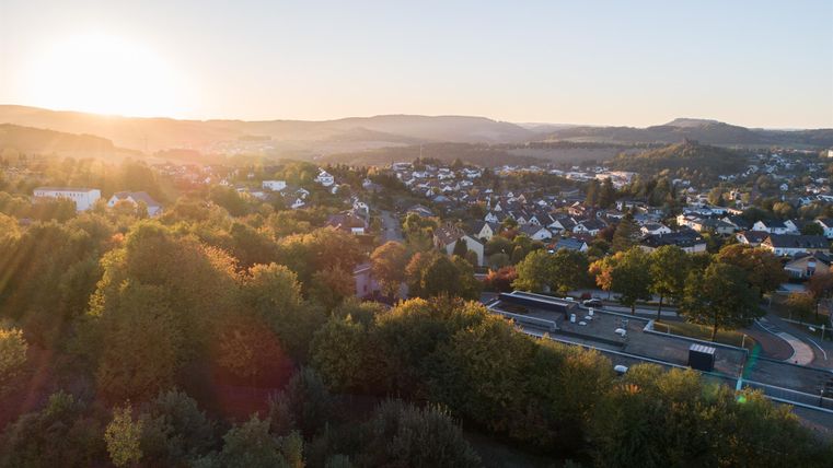 A picturesque landscape with houses and gentle hills in the background. The sun is rising in the background, bathing the scene in warm light.
