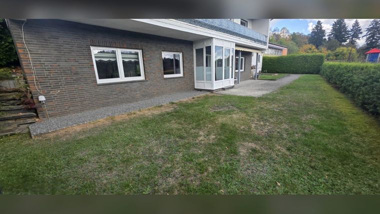Un jardin soigné avec de la pelouse et une terrasse attenant à une maison. À l'arrière-plan, on peut voir des buissons et des arbres.